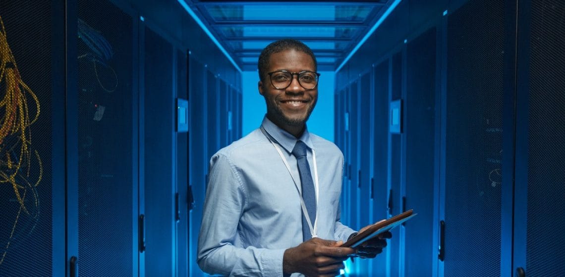 Waist up portrait of smiling African American man standing by server cabinet while working with supercomputer in data center and holding tablet, copy space